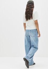 Hellblaue Schlaghose mit einem ausgewaschenen Finish, kombiniert mit einem weißen T-Shirt mit kurzen Ärmeln. Schwarze flache Schuhe, von hinten gezeigt.