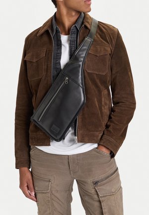 Uomo che indossa una giacca in suede marrone, borsa a tracolla in pelle nera, camicia bianca e pantaloni cargo beige con tasche con cerniera.