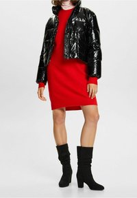 Czarna błyszcząca krótka kurtka puchowa na czerwonej sweterkowej sukience. Modelka ma na sobie czarne luźne botki na obcasie. Prosty, nowoczesny design.