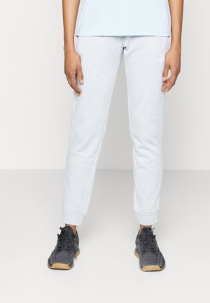 Person standing wearing light gray sweatpants and black athletic shoes with tan soles on a plain white background.