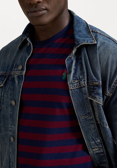 Τζάκετ από τζιν με μεταλλικά κουμπιά πάνω από μπλούζα φανέλα με navy και μπορντώ ρίγες που διαθέτει πράσινο λογότυπο. Το τζάκετ έχει φθαρμένες λεπτομέρειες.