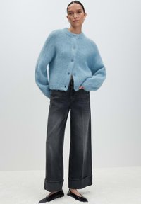 Donna che indossa un cardigan bottonato grigio chiaro e peloso, jeans grigi scuri a gamba larga con risvolti e ballerine nere a punta, in piedi su uno sfondo neutro.