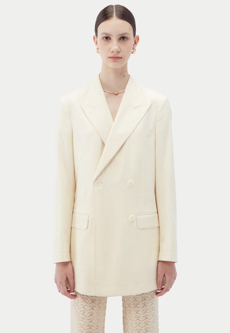 Blazer croisé en tissu crème clair, doté de revers crantés, deux poches avant, boutons de fermeture et coupe ajustée.