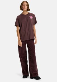 Bruine katoenen t-shirt met een roze bloemenontwerp op de linkerkant van de borst. Gepaard met wijde maroon corduroy broek. Zwarte schoenen zichtbaar.