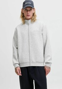 Ung mand med langt blondt hår iført en grå zip-up jakke, sorte bukser og en mørkeblå kasket med "JACK & JONES" logo.