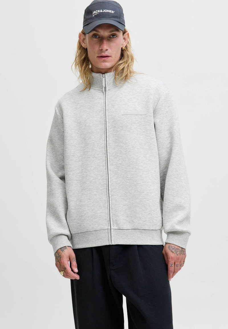 Joven con cabello largo y rubio que lleva una chaqueta gris con cremallera, pantalones negros y una gorra azul marino con el logo "JACK & JONES".