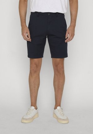 Homme portant un short bleu marine avec des poches latérales, des baskets blanches, et un t-shirt blanc, debout devant un fond clair uni.