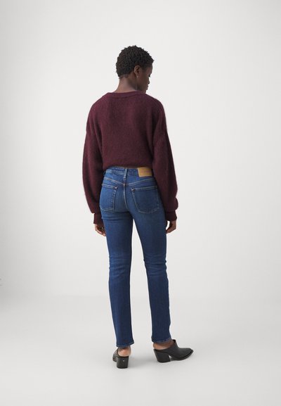 Suéter de punto burdeos con un corte holgado, combinado con vaqueros ajustados azules que tienen bolsillos traseros y un parche de cuero marrón, complementado con tacones negros.