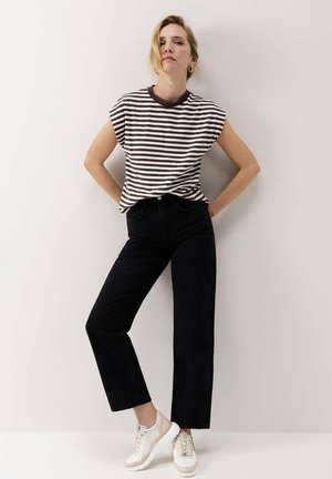 Mujer con una camiseta sin mangas de rayas negras y blancas, pantalones negros y zapatillas blancas, de pie con las manos en las caderas contra una pared lisa.