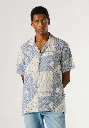 Jonge man die een korte mouwen blouse draagt met blauw en beige geometrische patronen, staand tegen een effen lichte achtergrond.