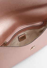 Sisustus roosansävyisessä synteettisessä käsilaukussa, jossa on sileä pinta, vetoketjullinen tasku ja "GUESS"-logotagi. Mukana kultaiset yksityiskohdat.