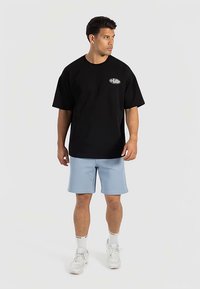 Schwarzes T-Shirt mit kurzen Ärmeln und einem kleinen Logo auf der Brust, kombiniert mit hellblauen Shorts und weißen Sneakers. Insgesamt schlichtes Design.