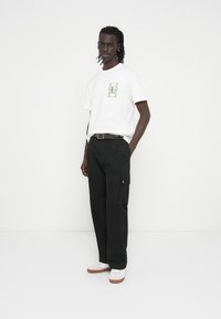 Άσπρο t-shirt με κοντά μανίκια και πράσινο λογότυπο, συνδυασμένο με μαύρο παντελόνι cargo με τσέπες και ασημένιο κουμπί, και λευκά αθλητικά παπούτσια.