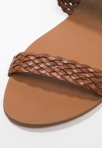 Sandalo in pelle intrecciata marrone con base piatta e una larga cintura. La cintura testurizzata presenta un design intrecciato su una suola liscia color beige.