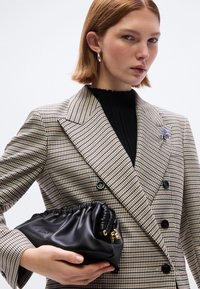 Δίπτυχος σακάκι με σχέδιο καρώ, μαύρο ζιβάγκο, κρατώντας ένα μαύρο δερμάτινο τσαντάκι χειρός με πτυχωτό πάνω μέρος και χρυσές μεταλλικές λεπτομέρειες.