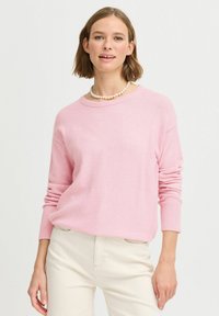 Světle růžový pletený svetr s kulatým výstřihem a žebrovanými manžetami, v kombinaci s béžovými kalhotami. Jednoduchý design, jemná textura a volný střih.