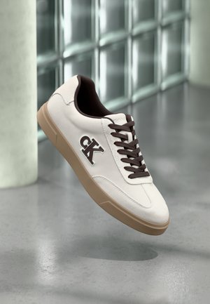 Biały sneaker z brązowymi akcentami, wykonany z zamszowego materiału, z płaskimi sznurówkami i gumową podeszwą. Na boku znajduje się logo, a powierzchnia ma gładką teksturę.