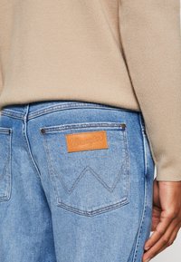 Modré džínsy Wrangler z denimové látky s hnědou koženou značkovou nášivkou, viditelné dva zadní kapsy, měděné nitě a detail cik-cak šití.