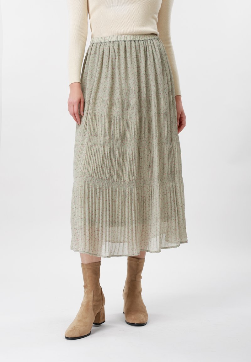 Žena oblečená v svetlozelenej plisovanej midi sukni s malým kvetinovým vzorom, béžovom rebrovanom svetri a hnedých semišových členkových topánkach s blokovým opätkom.