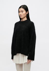 Mustast, lahtise lõikega kampsun, mis on valmistatud kohevast villast, pikkade varrukate ja kõrge kaelusega, kombineeritud plisseeritud heledat värvi seelikuga.