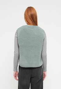 Colete de almofada em tecido verde suave com uma superfície macia e texturizada. Vista de trás mostra decote redondo e design sem mangas. Camisa cinzenta de manga comprida por baixo.