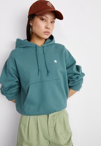 Teal hoodie met een voorzak en trekkoord, gecombineerd met een bruine pet met geborduurd logo. Model draagt lichtgroene shorts.