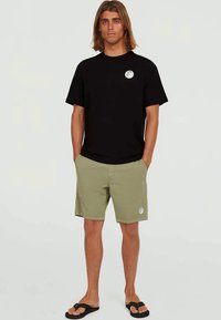 Young man standing with hands in pockets, wearing black t-shirt, olive green shorts, and black flip-flops against plain background.
