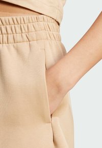 Joggers de algodón beige con cintura elástica, que cuentan con bolsillos laterales y una textura suave. Mano visible en un bolsillo.