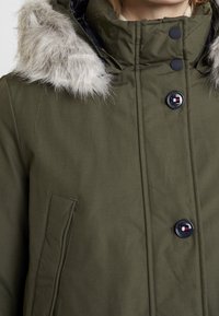 Olivově zelená parka bunda s kapucí lemovanou umělou kožešinou, předními knoflíky a detailem boční kapsy. Hladká textura se strukturovaným designem.