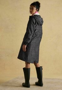 Vodoodolný čierny kabát s kvetinovým vzorom, so šnúrkovou kapucňou, elastickými manžetami a centrálnym ventilačným otvorom na chrbte, v kombinácii s čiernymi gumovými čižmami po kolená.