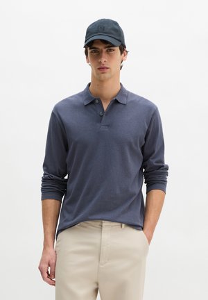 Junger Mann trägt ein dunkelblaues langärmeliges Polohemd, beigen Hosen und eine passende dunkelblaue Baseballkappe, steht mit einer Hand in der Tasche.