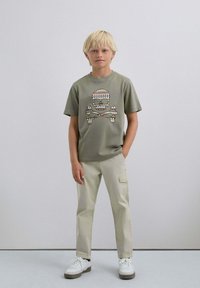 Camiseta verde oliva con un gráfico de calavera y tibias de colores. Pantalones cargo beige, zapatillas blancas con suelas grises. Fondo liso.