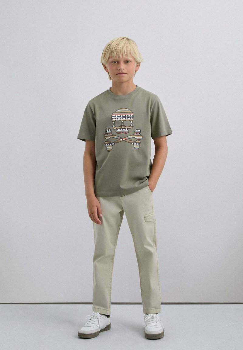 Camiseta verde oliva con un gráfico de calavera y tibias de colores. Pantalones cargo beige, zapatillas blancas con suelas grises. Fondo liso.