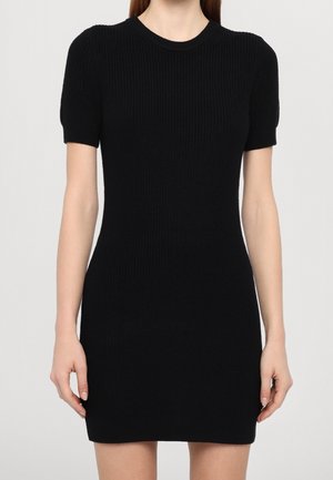 Kobieta ubrana w dopasowaną, czarną dzianinową sukienkę z krótkim rękawem i okrągłym dekoltem na jednolitym jasnym tle.