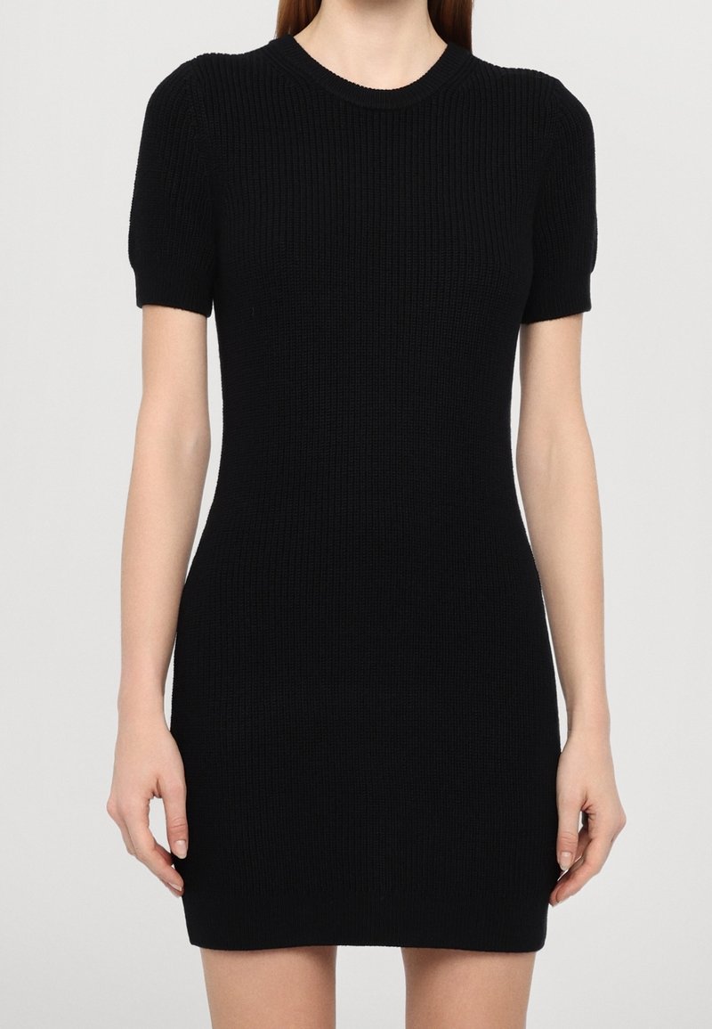 Femme portant une robe noire en maille moulante à manches courtes et col rond, sur un fond clair uni.