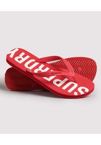 Tongs rouges avec le logo "SUPERDRY" en blanc, en matériau en caoutchouc, semelle intérieure texturée et semelle à motif en chevrons pour l'adhérence.