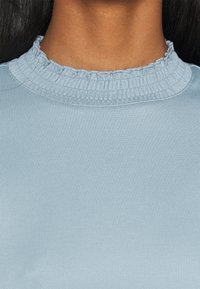 Top azzurro chiaro con una texture a coste e un colletto arricciato e volant. Il tessuto sembra morbido e leggermente elasticizzato.
