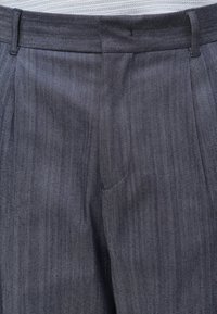 Plano aproximado da cintura e da zona do fecho de calças cinzentas pregueadas feitas de tecido texturizado com passadores para cinto.