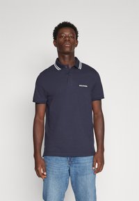Polo shirt blu navy con colletto a strisce bianche, maniche corte e logo ricamato sul petto. Indossato con jeans azzurri chiari.