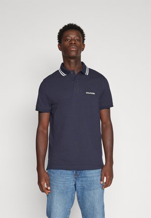 Polo shirt blu navy con colletto a strisce bianche, maniche corte e logo ricamato sul petto. Indossato con jeans azzurri chiari.