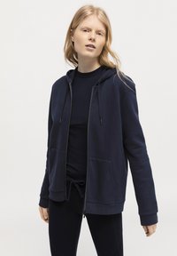 Marinblå zip-hoodie i mjukt tyg, med en kenguruficka, justerbar dragsko på huvan och ribbade ärmslut. Buren över en matchande topp.