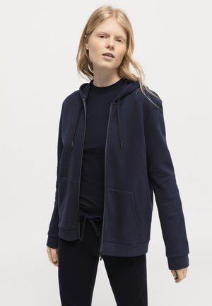 Sudadera con cremallera en color navy, hecha de una tela suave, que cuenta con un bolsillo tipo canguro, capucha ajustable con cordón y puños de canalé. Se lleva sobre una camiseta a juego.