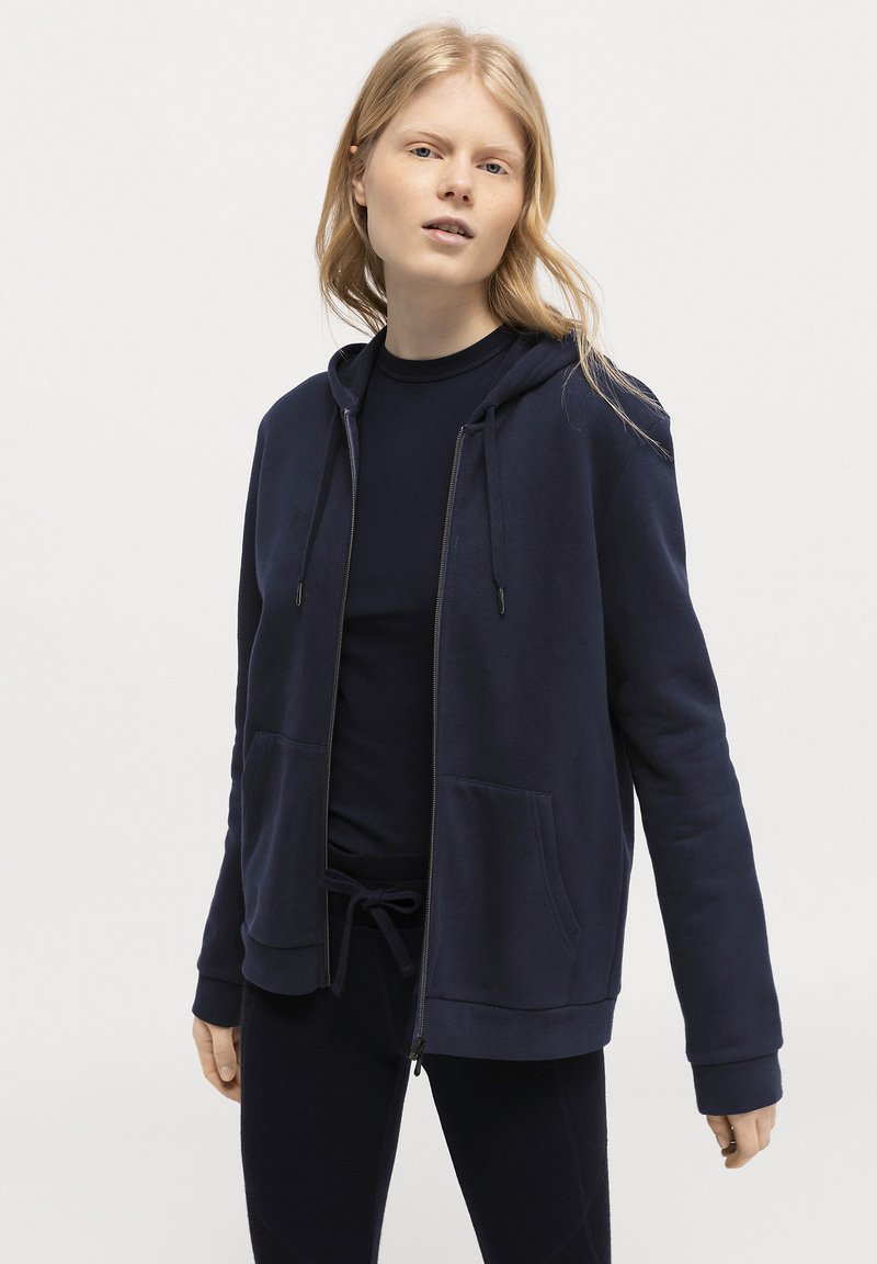 Marinblå zip-hoodie i mjukt tyg, med en kenguruficka, justerbar dragsko på huvan och ribbade ärmslut. Buren över en matchande topp.