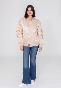 Jasno beżowa satynowa kurtka bomber z zamkiem z przodu, kieszeniami po bokach, ściągaczami na mankietach i pomarańczową metką na dolnym brzegu. Połączona z dzwonowymi dżinsami.