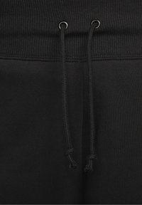 Černý elastický pas s vroubkovanou strukturou a dvěma černými šňůrkami uvázanými na koncích, procházejícími kovovými očky na černé látce.