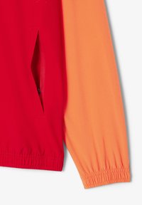 Giacca bloccata nei colori rosso e arancione con tasca con zip e polsini elasticizzati. Tessuto liscio con una texture leggera.