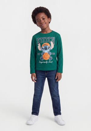 Garçon aux cheveux bouclés portant un t-shirt vert à manches longues avec Stitch tenant un ballon de basket, jean bleu et baskets blanches.