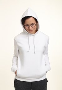 Hoodie bianco con cappuccio regolabile, maniche lunghe e tasche frontali. Texture liscia, design casual, indossato con pantaloni grigi.