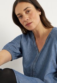 Lichtblauwe denim jurk met een V-hals, korte pofmouwen en geraffineerde details aan de voorkant, gecombineerd met zwarte leggins.