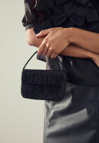 Woman holding a small black beaded handbag, wearing a sheer black top with bows and a black leather skirt.
